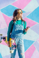Fototapeta premium Pretty woman wearing a green sweater laughing in front of a colorful background with smartphone and skateboard