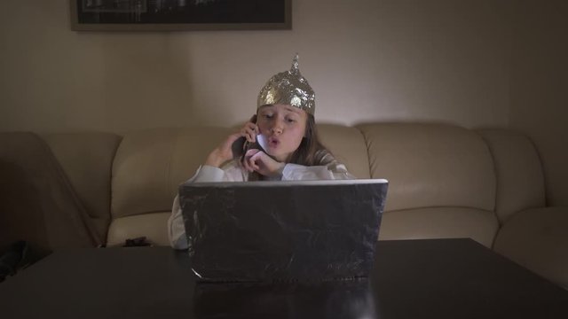 Young woman in Tin Foil Hat speaks expressively on the phone, waving hands. Foil hat protects her from 5G waves, electromagnetic fields, mind control, mind reading, global conspiracy