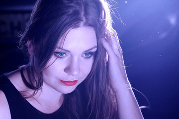 Young beautiful female with green eyes and red lipstick looks down, smiling. Night club or youngsters party, dark electric blue background, back light.