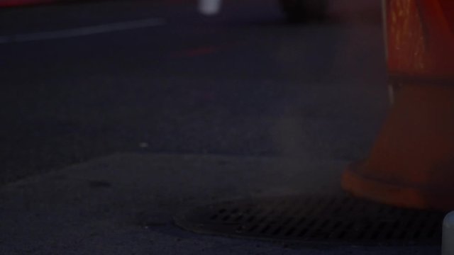 Smoking manhole cover near construction cone marker, New York City street with cars passing by, evening shot