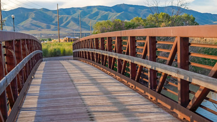 Fototapeta premium Pano Sunlit wooden deck and metail railing of bridge over lake with mountain view
