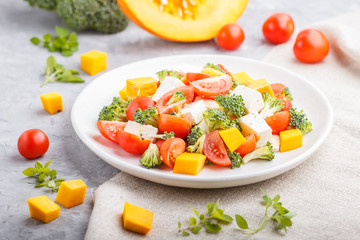 Vegetarian salad with broccoli, tomatoes, feta cheese, and pumpkin on white ceramic plate on a gray concrete background, side view, close up.