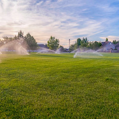 Square frame Sprinklers watering green grassy field with homes and cloudy blue sky background