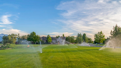 Pano frame Sprinklers spraying water on green grasses with homes mountain and blue sky view - obrazy, fototapety, plakaty