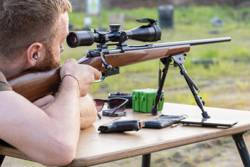 A man with a beard looks into the scope of a sniper rifle, prepares to shoot