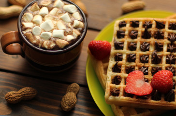 Breakfast. Waffles filled with chocolate cream, decorated with strawbierries hearts. Cup of coffee with mini marshmallows. Peanuts around. On wooden table.