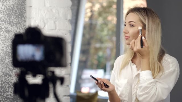 A makeup artist is applying highlighter with a brush on her face. The pretty blond is commenting the makeup tutorial on while filming with a camera.