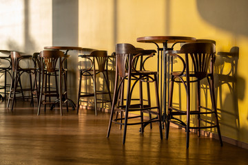 Tall vintage wooden tables and chairs stand in a cafe. A soft shadow from sunlight is reflected in the foreground on the wall and floor, in the background is a cold shadow. Bottom side view