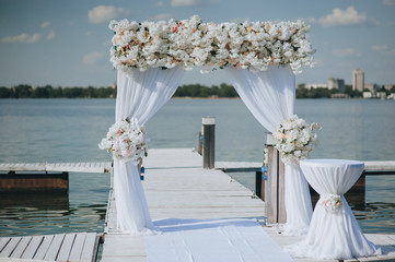 A wedding arch made of white fabric and hydrangea flowers stands on a pier, amid a river and sea....