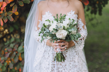The bride in a white lace dress holds in her hands a wedding bouquet of roses. Closeup portrait of fresh flowers on the nature in autumn. Photography, concept.
