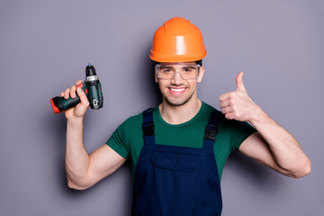 Portrait of handsome man repairer successful handyman hold perforator show thumb up sign wear orange hard hat green t-shirt protective goggles isolated over grey color background