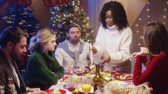 Pretty nice mood african girl carving hot turkey and treating her friends with delicious food, celebrating winter dinner party together on Christmas Eve.