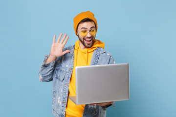 Joyful young hipster guy in fashion jeans denim clothes posing isolated on pastel blue background. People lifestyle concept. Mock up copy space. Working on laptop pc computer, waving hand, greeting.