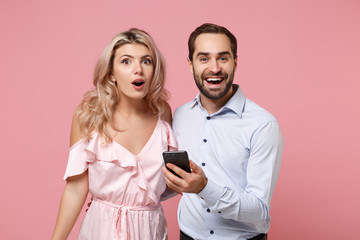 Shocked young couple two guy girl in party outfit celebrating isolated on pastel pink background. Valentine's Day Women's Day birthday holiday party concept. Using mobile phone, typing sms message.