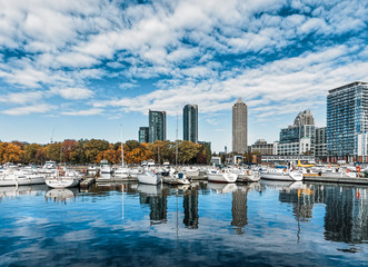Fototapeta na wymiar Sailboats are moored in the Toronto harbour near the Toronto Naval Divison building are reflected in the water on a bright sunny fall day with changing foliage, the Toronto skyline