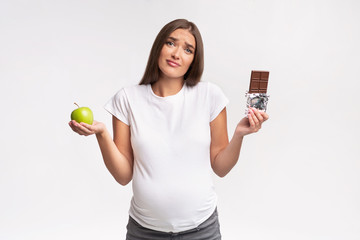 Confused Pregnant Lady Holding Apple And Chocolate Standing In Studio