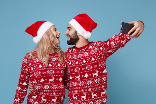 Funny young couple guy girl in Christmas sweater Santa hat posing isolated on blue background. New Year 2020 celebration holiday party concept. Mock up copy space. Doing selfie shot on mobile phone.