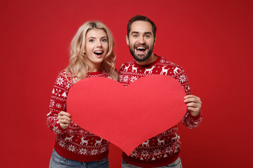 Excited young couple guy girl in Christmas knitted sweaters posing isolated on red background. Happy New Year 2020 celebration holiday party concept. Mock up copy space. Hold empty blank red heart.