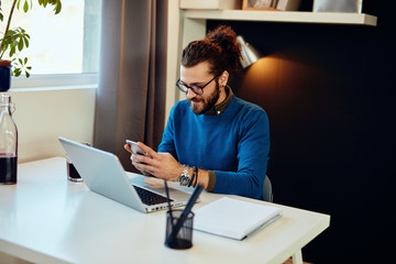 Smiling caucasian hipster sitting in his office and using smart phone for responding an e-mail.