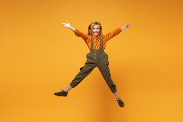 Fototapeta na wymiar Smiling little kid girl 12-13 years old in turtleneck, jumpsuit isolated on orange yellow wall background in studio. Childhood lifestyle concept. Mock up copy space. Jumping, spreading hands and legs.