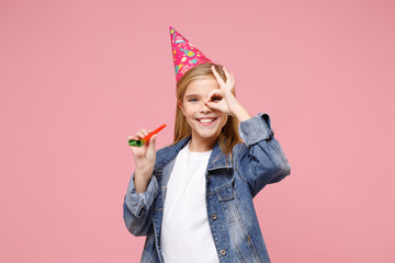 Smiling little blonde kid girl 12-13 years old in denim jacket birthday hat isolated on pastel pink background. Childhood lifestyle concept. Mock up copy space. Hold party pipe keeping hand near eyes.