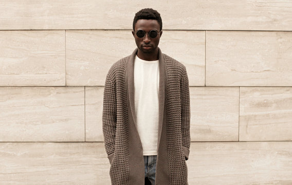 Stylish african man wearing brown knitted cardigan, sunglasses, male model posing on city street over brick wall background