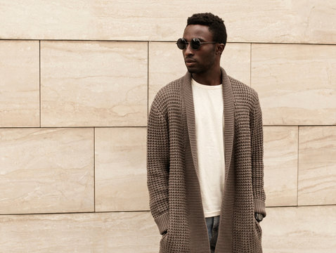 Stylish african man model looking away wearing brown knitted cardigan, sunglasses on city street over brick wall background