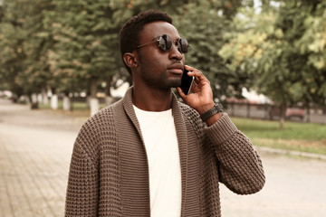 Portrait african man calling on smartphone wearing brown knitted cardigan and sunglasses on city...