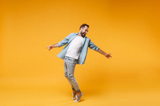 Laughing young bearded man in casual blue shirt posing isolated on yellow orange background studio portrait. People emotions lifestyle concept. Mock up copy space. Standing on toes, spreading hands.