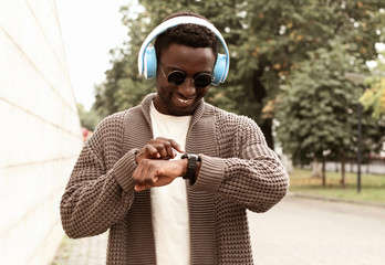 Portrait smiling african man looking at smart watch using voice assistant or takes calling in wireless headphones on city street