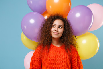 Pensive young african american girl in orange knitted clothes isolated on pastel blue background with colorful air balloons. Birthday holiday party people emotions concept. Celebrating, looking up.