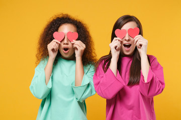 Two excited women friends european african american in pink green clothes posing isolated on yellow...