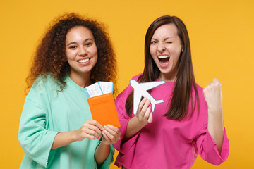 Two women friends european american girls in pink green clothes posing isolated on yellow background. People lifestyle concept. Mock up copy space. Hold passport ticket airplane doing winner gesture.