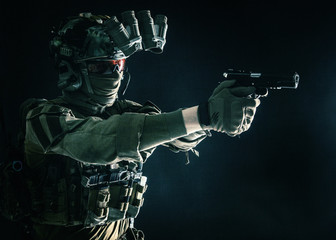 Modern soldier, counter terrorist squad fighter in combat uniform, helmet and tactical radio headset, aiming service pistol in darkness, shooting with handgun, studio portrait on dark background