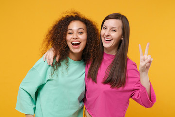 Two cheerful women friends european and african american girls in pink green clothes posing isolated on yellow background. People lifestyle concept. Mock up copy space. Hugging, showing victory sign.