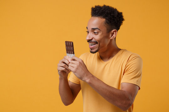 Joyful young african american guy in casual t-shirt posing isolated on yellow orange background studio portrait. People sincere emotions lifestyle concept. Mock up copy space. Holding chocolate bar.