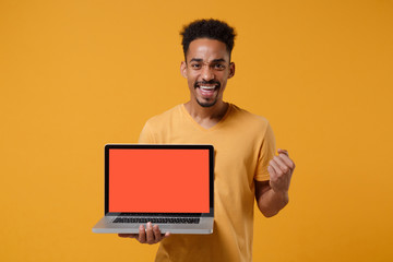 Young african american guy in t-shirt posing isolated on yellow orange background. People lifestyle concept. Mock up copy space. Hold laptop pc computer with blank empty screen, doing winner gesture.