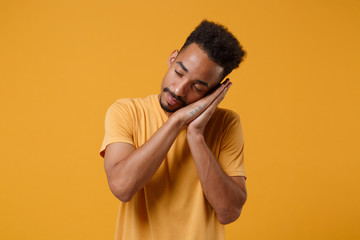 Relaxed young african american guy in casual t-shirt posing isolated on yellow orange background studio portrait. People lifestyle concept. Mock up copy space. Sleep with folded hands under cheek.