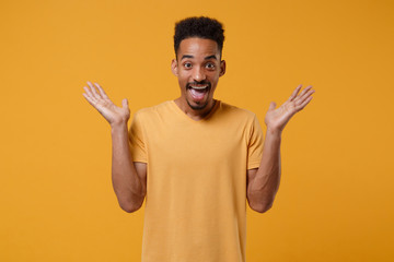 Fototapeta premium Excited young african american guy in casual t-shirt posing isolated on yellow orange wall background studio portrait. People lifestyle concept. Mock up copy space. Spreading hands keeping mouth open.