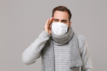 Young man in gray sweater, scarf isolated on grey background. Healthy lifestyle, ill sick disease treatment, cold season concept. Mock up copy space. Wearing sterile face mask put hand on temple head.