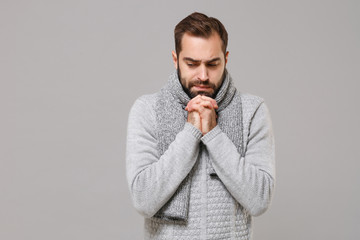 Young man in gray sweater, scarf posing isolated on grey background, studio portrait. Healthy fashion lifestyle, people emotions, cold season concept. Mock up copy space. Hold hands folded in prayer.