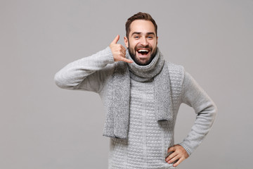 Funny young man in gray sweater scarf posing isolated on grey background studio portrait. Healthy fashion lifestyle cold season concept. Mock up copy space. Doing phone gesture like says call me back.