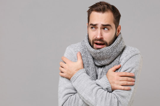 Frozen young man in gray sweater posing isolated on grey wall background, studio portrait. Healthy fashion lifestyle, cold season concept. Mock up copy space. Holding hands folded, huging himself.