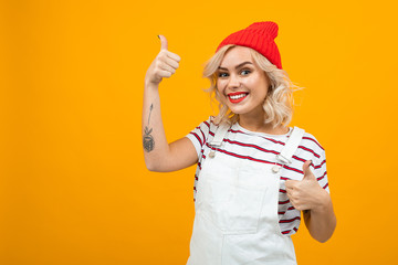 Fototapeta na wymiar Beautiful young woman with short blonde curly hair and bright makeup in white overalls and red hat likes something and smiles, portrait isolated on orange background