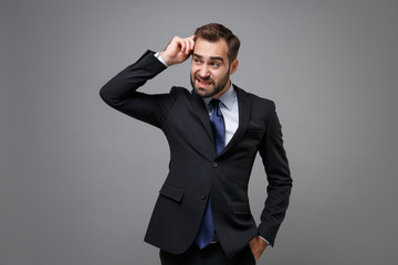 Concerned puzzled young bearded business man in classic black suit shirt tie posing isolated on grey background. Achievement career wealth business concept. Mock up copy space. Putting hand on head.