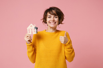 Smiling young brunette woman girl in yellow sweater posing isolated on pastel pink background studio portrait. People lifestyle concept. Mock up copy space. Hold house bunch of keys, showing thumb up.
