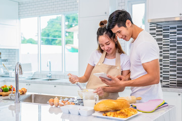 Obraz na płótnie Canvas Happy Asian couples cooking and baking cake together in kitchen room. Man and woman looking to tablet follow recipe step at home. Love and happiness concept. Sweet honeymoon and Valentine day theme