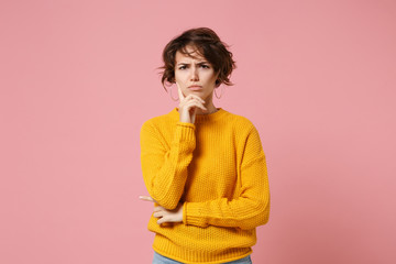 Displeased pensive young brunette woman girl in yellow sweater posing isolated on pastel pink wall background studio portrait. People lifestyle concept. Mock up copy space. Put hand prop up on chin.