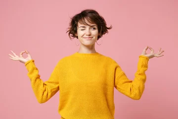 Foto op Canvas Grappige jonge brunette vrouw meisje in gele trui poseren geïsoleerd op pastel roze achtergrond. Mensen levensstijl concept. Bespotten kopie ruimte. Houd handen vast in yogagebaar ontspannen mediteren opzij kijken. © ViDi Studio