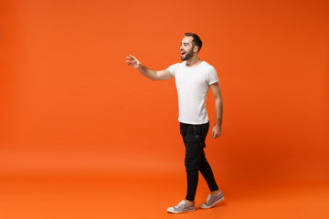 Funny young man in casual white t-shirt posing isolated on bright orange wall background studio portrait. People sincere emotions lifestyle concept. Mock up copy space. Stand with outstretched hand.